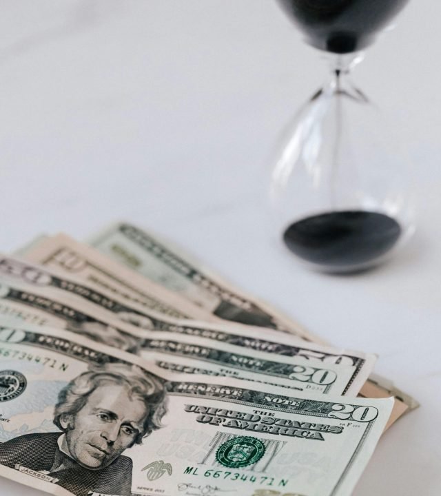 From above of transparent hourglass with black sand and heap of American dollars with image of president